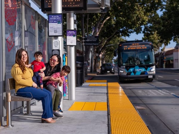 Two women and two young children wait at a bus station on a tree lined street for an approaching bus